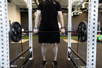 Lifter performing deadlifts inside the rack using a bar loaded with a pair of 45lb plates and also black short resistance bands attached via band pegs for added resistance.